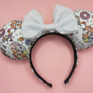 May include: A headband featuring two round ears with a white background patterned with cartoon characters, including a fox and a rabbit. A large, white, sequined bow is centered between the ears. The headband has a black interior lining.