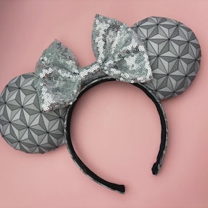 May include: A headband featuring two circular ears with a grey and black geometric triangular pattern. A large, central bow covered in shimmering silver sequins sits between the ears. The headband frame is black and has a smooth, rounded shape.