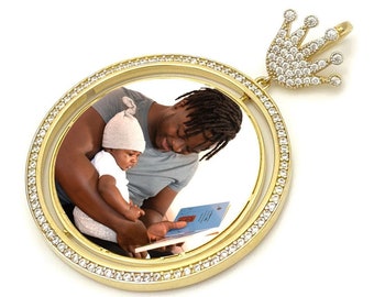 May include: Gold pendant with a round photo frame featuring a father and child reading a book. The frame is surrounded by a row of clear cubic zirconia stones. The pendant is topped with a crown design also adorned with clear cubic zirconia stones.