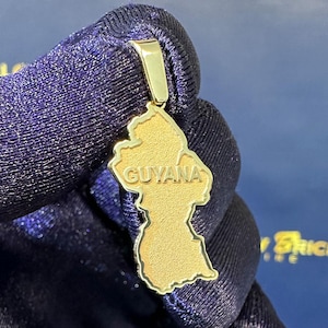 May include: A gold-colored pendant in the shape of Guyana, with the country's name engraved on it. The pendant is suspended from a gold bail. The background is a dark blue fabric.