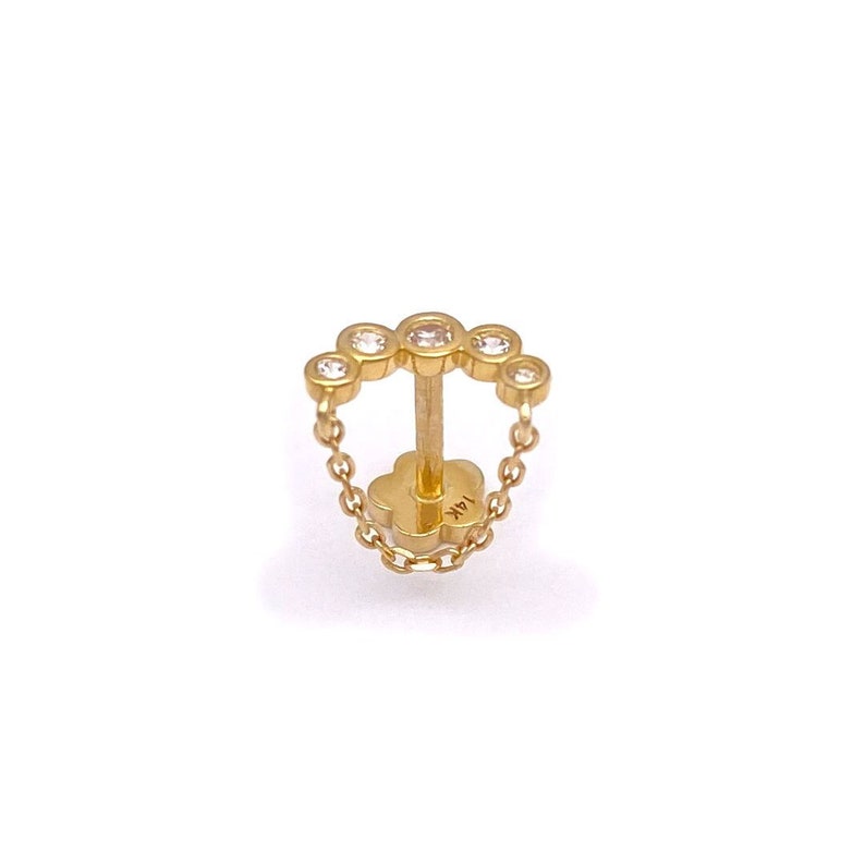 May include: A gold earring with a chain and five clear gemstones in a circular design. The earring has a post and a flower-shaped backing. The metal is stamped with "14K".