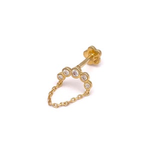 May include: A gold-colored earring with a chain and a series of clear, round gemstones. The earring features a flower-shaped backing and a chain that dangles in a semi-circle. The metal is shiny and reflective.