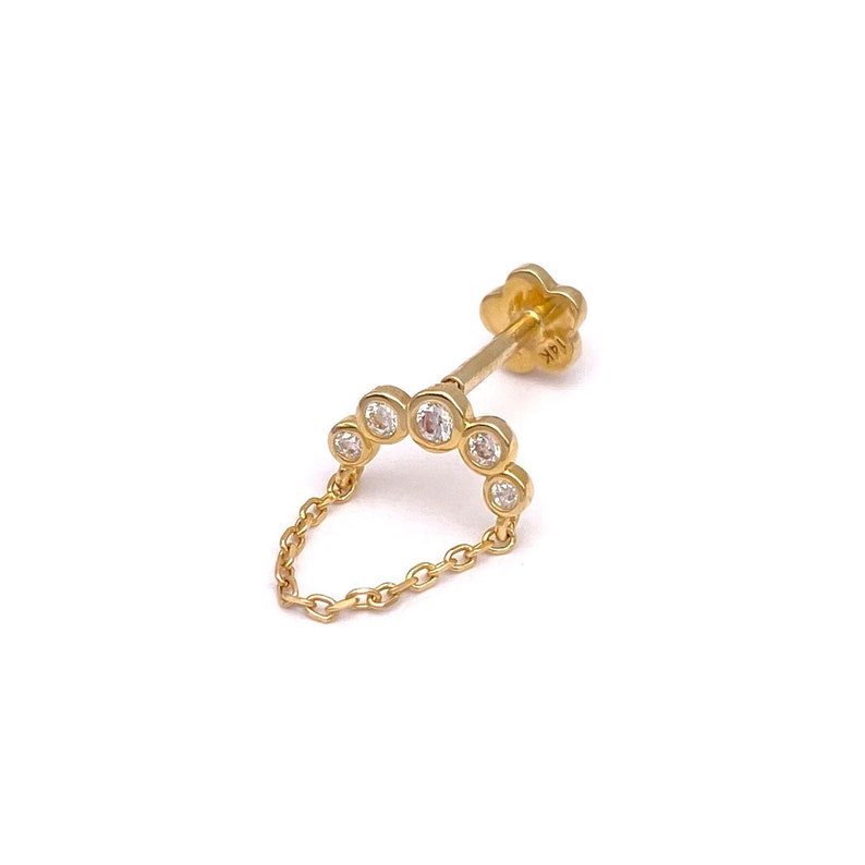 May include: A gold-colored earring with a chain and a series of clear, round gemstones. The earring features a flower-shaped backing and a chain that dangles in a semi-circle. The metal is shiny and reflective.