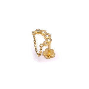 May include: A gold-colored earring with a chain and multiple clear, round gemstones. The earring has a flower-shaped base and is set against a white background. The chain connects the gemstones to the base.