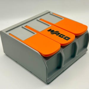 May include: A grey and orange electrical connector with the word "WAGO" printed in black. The connector has three orange levers and is designed for electrical wiring applications.
