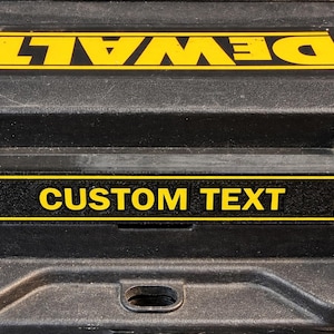 Könnte beinhalten: Schwarze Werkzeugkiste mit gelbem "DEWALT"-Logo und einem gelb-schwarzen Etikett mit der Aufschrift "CUSTOM TEXT". Die Werkzeugkiste besteht aus einem strapazierfähigen Material und ist zur Aufbewahrung von Werkzeugen konzipiert.