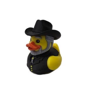 Op de afbeelding: Een gele rubberen eend verkleed als cowboy, met een zwarte hoed, een zwart vest met drie kleine knoopjes en een grijze baard. De eend heeft een oranje snavel en een bolo-das accessoire om de nek, tegen een effen witte achtergrond.