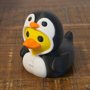 Op de afbeelding: Een gele rubberen eend in een zwart-wit pinguïnkostuum. Het kostuum heeft een capuchon met een pinguïngezicht, zwarte vleugels aan de zijkanten en een wit buikgedeelte. De eend heeft een feloranje snavel en zwarte ogen die door de capuchon zichtbaar zijn.