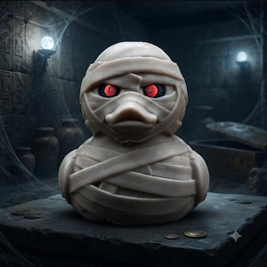 Puede incluir: Una figura de pato de goma envuelta en vendas de color beige para parecer una momia. El pato presenta ojos rojos brillantes situados dentro de las capas de vendas, creando un contraste marcado con el material claro de las mismas.