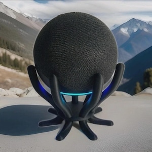 May include: A black, spherical smart speaker sits in a black, stylized stand with curved arms and a circular base. The stand has a blue light at the base. The background shows a mountain range.