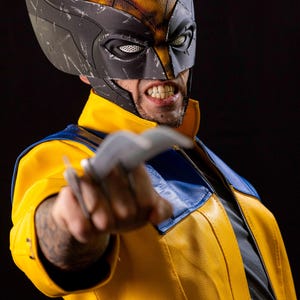 May include: A person wearing a yellow and blue leather jacket with a black and gray mask. The person is holding a silver claw in their right hand. The person is looking at the camera with a fierce expression.
