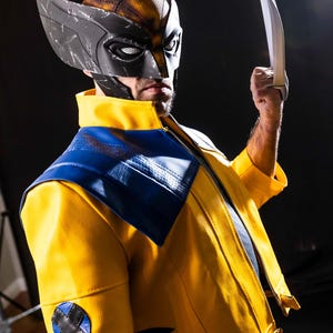 May include: A person wearing a yellow and blue jacket with a black and gray mask and holding a silver claw. The mask has a textured surface and the jacket has a black X on the left sleeve.
