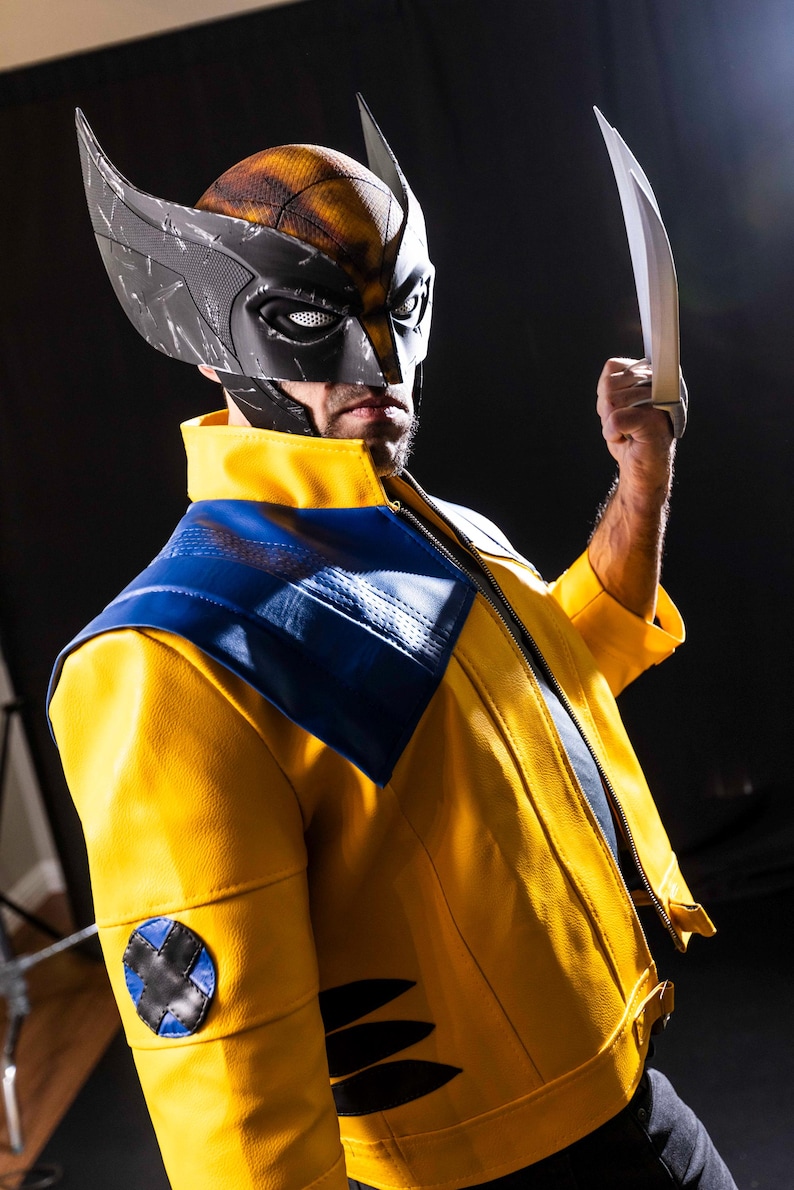 May include: A person wearing a yellow and blue jacket with a black and gray mask and holding a silver claw. The mask has a textured surface and the jacket has a black X on the left sleeve.