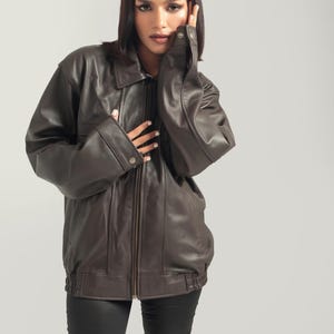May include: A brown leather jacket with a zipper closure and a stand-up collar. The jacket has a relaxed fit and is worn by a person with dark hair.