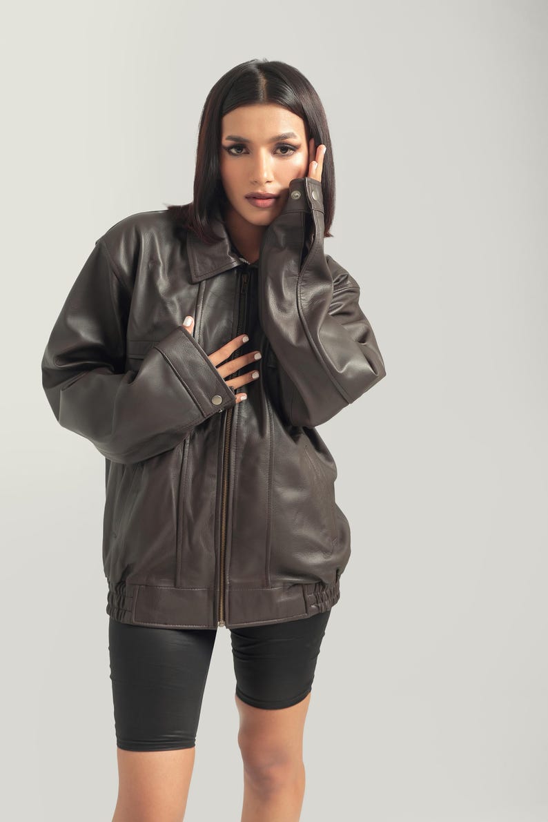 May include: A brown leather jacket with a zipper closure and a stand-up collar. The jacket has a relaxed fit and is worn by a person with dark hair.