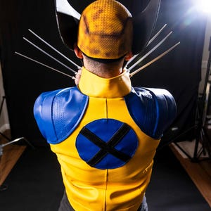 May include: A person wearing a yellow and blue costume with a yellow helmet and black claws. The costume has a blue circle with a black X on the back.