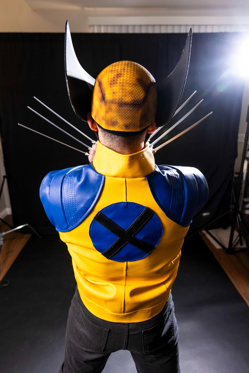 May include: A person wearing a yellow and blue costume with a yellow helmet and black claws. The costume has a blue circle with a black X on the back.