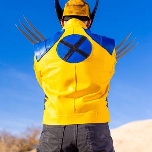 May include: A yellow leather jacket with a blue circle on the back featuring a black X. The jacket has a blue collar and blue accents on the shoulders. The person is wearing black jeans.