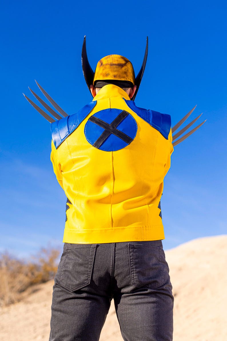 May include: A yellow leather jacket with a blue circle on the back featuring a black X. The jacket has a blue collar and blue accents on the shoulders. The person is wearing black jeans.