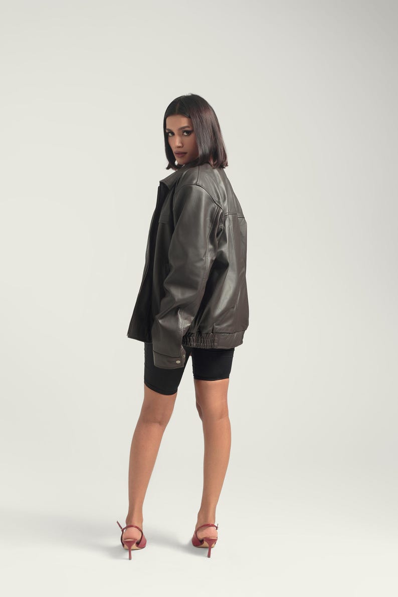 May include: A woman wearing a brown leather jacket, black biker shorts, and burgundy heeled shoes. She is standing with her back to the camera and looking over her shoulder.
