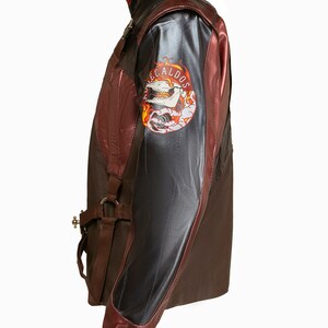 Puede incluir: Una chaqueta de cuero marr&oacute;n con un panel de cuero negro en el lado izquierdo del pecho y un panel de cuero negro en la manga derecha. La chaqueta tiene una cremallera y un cuello de punto marr&oacute;n. La manga derecha tiene un parche con una calavera y llamas y el texto "NE CALDOS".