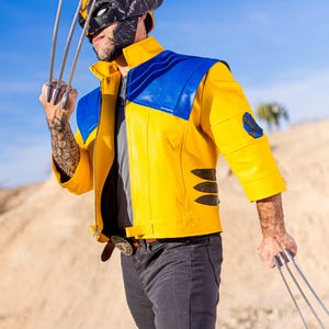 May include: A person wearing a yellow and blue leather jacket with a black and gray mask, holding silver claws in each hand. The person is standing in a desert landscape.