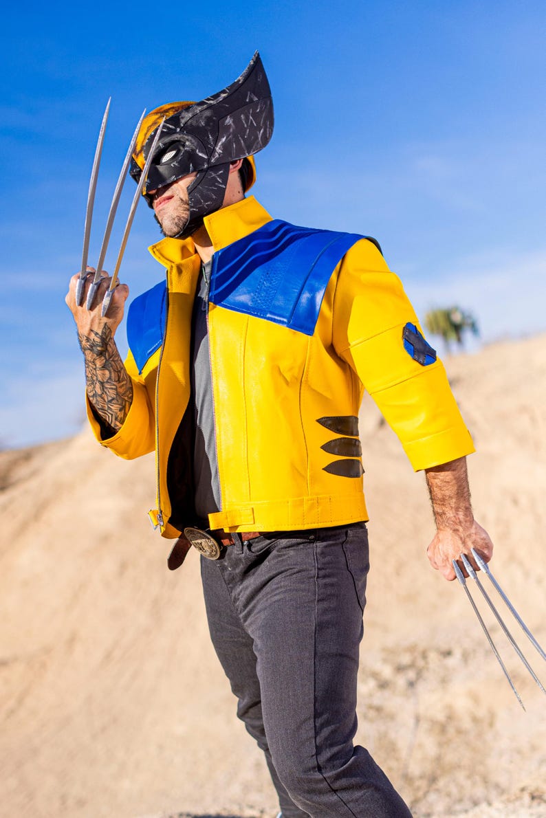 May include: A person wearing a yellow and blue leather jacket with a black and gray mask, holding silver claws in each hand. The person is standing in a desert landscape.