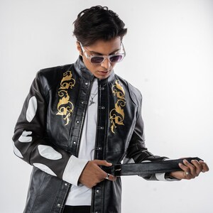 May include: A black leather jacket with gold embroidery and white accents. The jacket has a button-up front and a zipper closure. The model is wearing a white t-shirt and black pants.