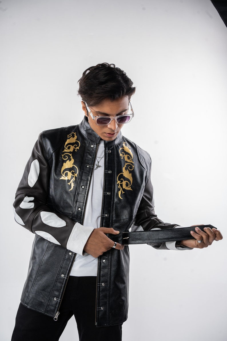 May include: A black leather jacket with gold embroidery and white accents. The jacket has a button-up front and a zipper closure. The model is wearing a white t-shirt and black pants.