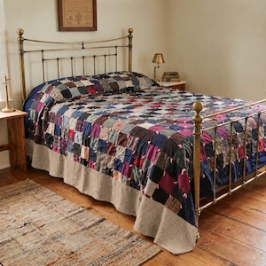 May include: A patchwork quilt featuring a grid of multi-colored fabric squares in shades of blue, red, purple, and brown, accented with small pink fabric ties at each corner, draped over a brass metal bed frame with a solid beige bed skirt.