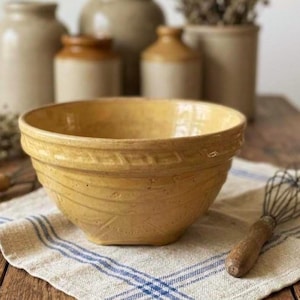 May include: A yellow ceramic mixing bowl with a wide rim and a textured geometric pattern around the upper exterior. The bowl has a smooth, glazed finish and a deep, rounded shape, positioned on a white and blue striped fabric cloth.