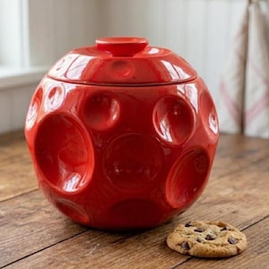 May include: A spherical, bright red ceramic cookie jar with a matching lid, featuring a textured surface covered in circular indentations of varying sizes. A single round chocolate chip cookie sits on the surface next to the jar.