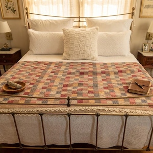 May include: A bed with a metal frame, adorned with white bedding and a patchwork quilt in shades of red, beige, and blue. A decorative pillow sits atop the bed, and a wooden bowl and journal are placed on the quilt.