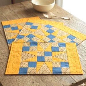 May include: A rectangular quilted placemat featuring a central star pattern composed of yellow, blue, and floral-print fabric squares. The border is solid yellow with a stitched leaf motif, and the design is repeated on a second placemat underneath.