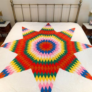 May include: A large quilt featuring a central star pattern composed of diamond-shaped fabric patches in vibrant colors including red, orange, yellow, green, blue, and pink, set against a solid white background fabric that covers the bed.
