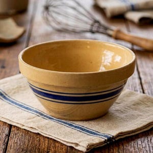 May include: A round, yellow ceramic mixing bowl featuring two horizontal dark blue stripes encircling the exterior. The bowl has a smooth, glazed finish and a wide, thick rim, presenting a classic design for kitchen preparation and food mixing.