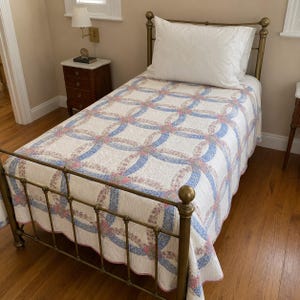 May include: A twin-sized bed with a brass metal frame featuring a white quilt with a blue and pink floral double wedding ring pattern. A single white pillow rests at the head of the bed, which is covered by the patterned quilt with scalloped edges.