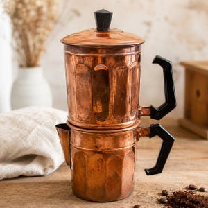 May include: A copper coffee maker featuring a two-part cylindrical body with vertical fluted detailing. It includes two black angular handles, a matching black hexagonal knob on the lid, and a small pouring spout on the lower section.