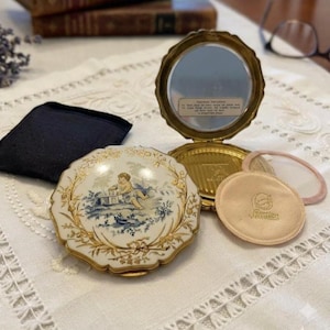 May include: A round gold-tone metal compact mirror with a scalloped edge and a white lid featuring a blue and gold illustration of a cherub. The interior includes a mirror with a label, a gold-tone powder well, a beige powder puff, and a black pouch.