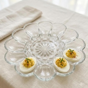 May include: A clear glass deviled egg serving platter with a scalloped edge and a central starburst pattern. The platter features twelve oval-shaped indentations, two of which contain halved hard-boiled eggs topped with yellow filling.