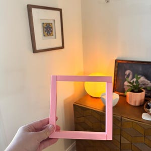 K&ouml;nnte beinhalten: Ein quadratischer, hellrosa Bilderrahmen wird vor einer Wand gehalten. Der Rahmen ist leer, ohne Bild. Der Hintergrund umfasst gerahmte Kunstwerke, dekorative Haken und einen Holzschrank.