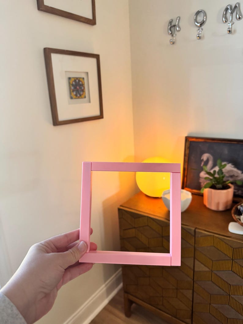K&ouml;nnte beinhalten: Ein quadratischer, hellrosa Bilderrahmen wird vor einer Wand gehalten. Der Rahmen ist leer, ohne Bild. Der Hintergrund umfasst gerahmte Kunstwerke, dekorative Haken und einen Holzschrank.