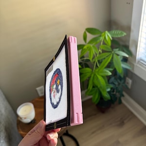 K&ouml;nnte beinhalten: Ein gerahmtes Nadelbild mit einer Zeichentrickfigur in einem blauen Kreis mit rotem und blauem Hintergrund. Der Rahmen ist schwarz mit einer rosa Seite.