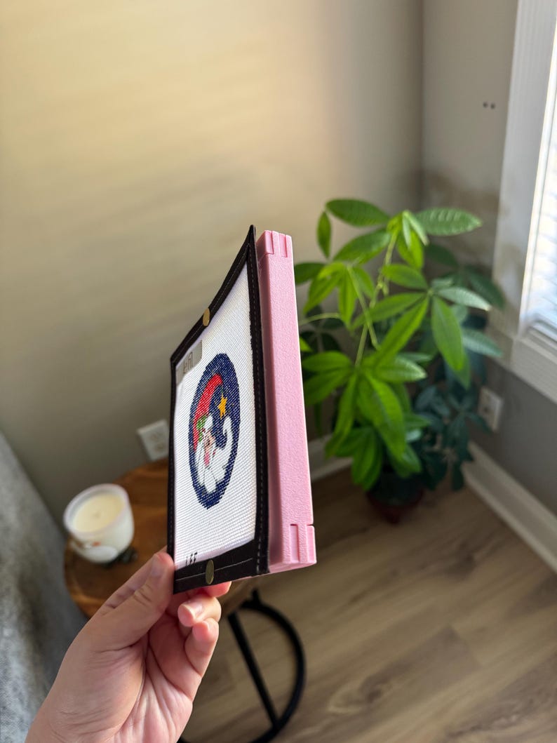 K&ouml;nnte beinhalten: Ein gerahmtes Nadelbild mit einer Zeichentrickfigur in einem blauen Kreis mit rotem und blauem Hintergrund. Der Rahmen ist schwarz mit einer rosa Seite.
