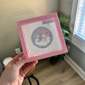 K&ouml;nnte beinhalten: Ein rosa Stickrahmen mit einem wei&szlig;en Netzmittelpunkt. Das Netz zeigt ein farbenfrohes, kreisf&ouml;rmiges Design. Der Rahmen wird von einer Hand gehalten, mit einer Uhr am Handgelenk. Der Hintergrund umfasst ein Fenster und einen Holzboden.
