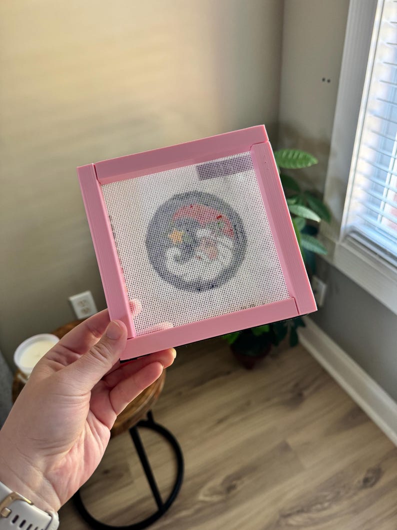 K&ouml;nnte beinhalten: Ein rosa Stickrahmen mit einem wei&szlig;en Netzmittelpunkt. Das Netz zeigt ein farbenfrohes, kreisf&ouml;rmiges Design. Der Rahmen wird von einer Hand gehalten, mit einer Uhr am Handgelenk. Der Hintergrund umfasst ein Fenster und einen Holzboden.