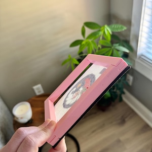 K&ouml;nnte beinhalten: Ein rosa Stickrahmen, der ein fertiges Kreuzstichdesign h&auml;lt. Der Rahmen ist abgewinkelt und zeigt das Kreuzstichmuster mit roten, wei&szlig;en und blauen Farben. Der Rahmen hat eine schwarze R&uuml;ckseite.
