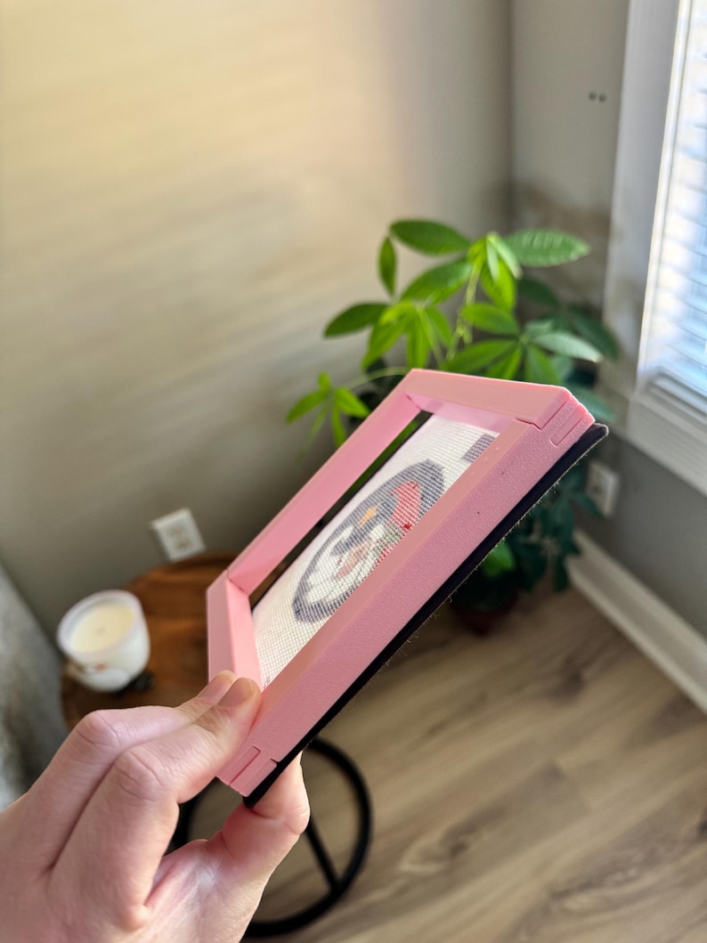 K&ouml;nnte beinhalten: Ein rosa Stickrahmen, der ein fertiges Kreuzstichdesign h&auml;lt. Der Rahmen ist abgewinkelt und zeigt das Kreuzstichmuster mit roten, wei&szlig;en und blauen Farben. Der Rahmen hat eine schwarze R&uuml;ckseite.