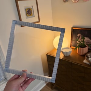 K&ouml;nnte beinhalten: Ein hellgrauer, quadratischer Bilderrahmen mit strukturierter Oberfl&auml;che. Der Rahmen wird vor einer Wand mit einem gerahmten Bild und dekorativen Gegenst&auml;nden gehalten. Der Rahmen ist f&uuml;r ein quadratisches Bild konzipiert.