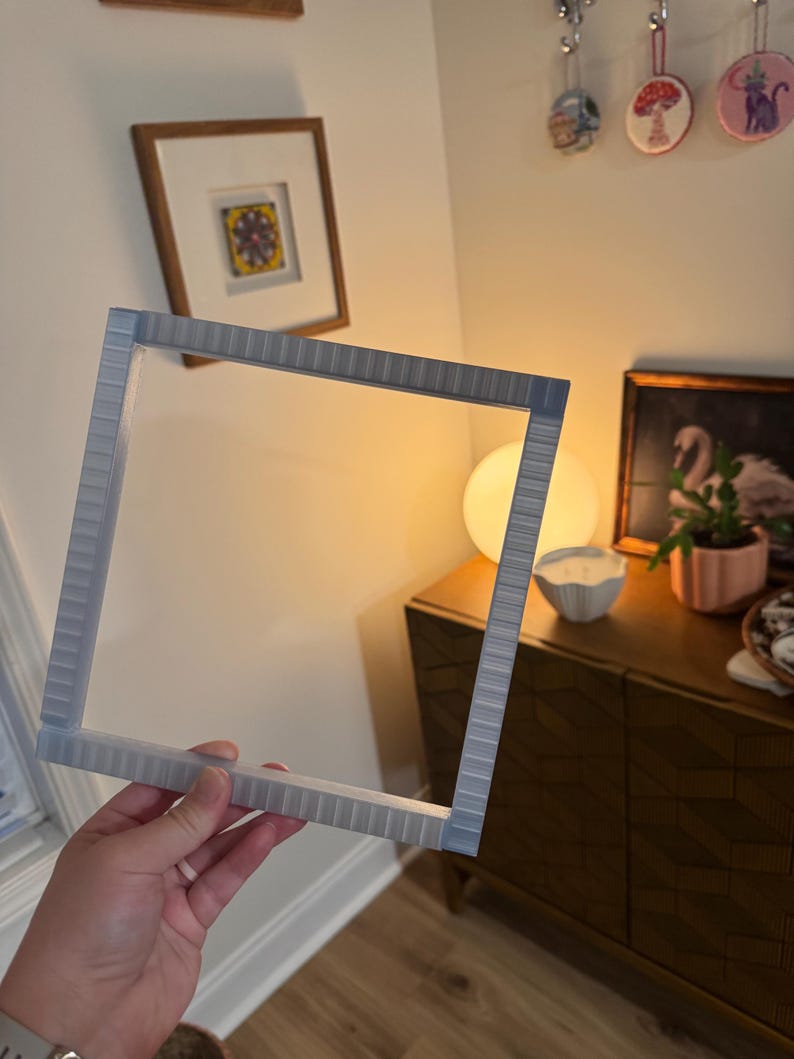 K&ouml;nnte beinhalten: Ein hellgrauer, quadratischer Bilderrahmen mit strukturierter Oberfl&auml;che. Der Rahmen wird vor einer Wand mit einem gerahmten Bild und dekorativen Gegenst&auml;nden gehalten. Der Rahmen ist f&uuml;r ein quadratisches Bild konzipiert.